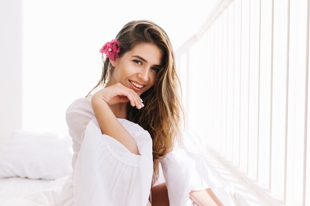 Chica encantadora alegre con increíble sonrisa y peinado romántico con mucho gusto posando en la sala blanca. Retrato de mujer joven linda con flor rosa en el pelo, sentada en la cama en la mañana soleada y riendo