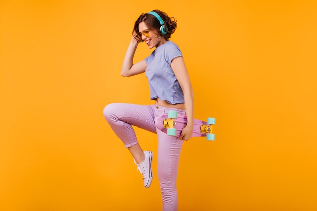 Chica encantadora delgada en auriculares azules bailando con una sonrisa. Modelo femenino bien proporcionado con longboard saltando con expresión de cara feliz.