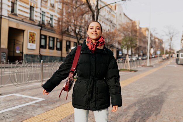 Una chica encantadora y emocionada con un maquillaje brillante y un peinado oscuro con una chaqueta oscura y una bufanda roja se está divirtiendo al aire libre en una calle soleada