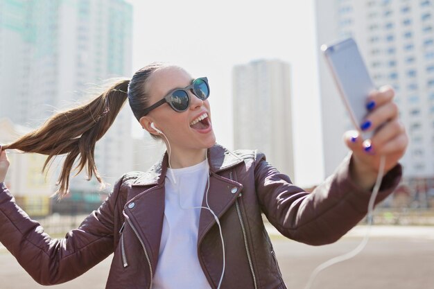 Foto gratuita una chica encantadora emocionada con el pelo recogido con gafas de sol y una chaqueta de cuero está haciendo selfie en un smartphone con verdaderas emociones felices