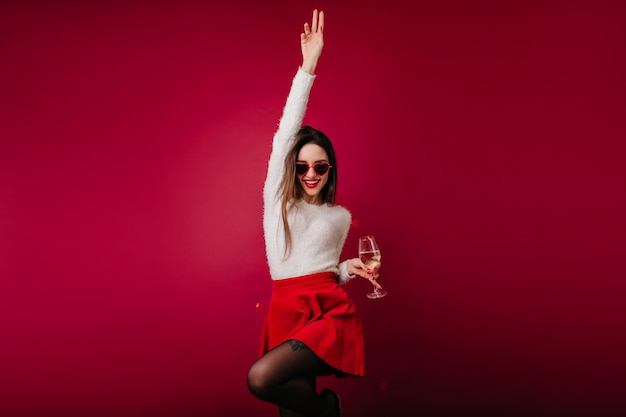 Chica encantadora en falda roja corta divertida bailando con copa de vino en la mano
