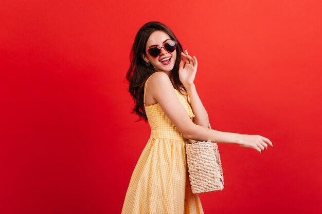 Chica encantadora de moda en vestido a cuadros riendo en la pared roja. Foto de modelo de mujer con gafas en forma de corazón y bolso de mimbre.