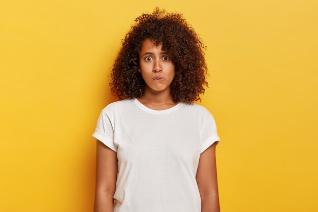 Una chica encantadora preocupada con el pelo tupido y rizado se muerde los labios, tiene una expresión nerviosa de sorpresa, se siente avergonzada de hablar en un lugar público y usa poses de ropa blanca sobre una pared amarilla. Concepto de emociones humanas