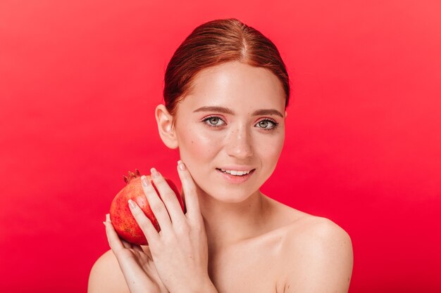 Chica encantadora sosteniendo granate con una sonrisa suave. Disparo de estudio de increíble dama de jengibre con frutas aisladas sobre fondo rojo.