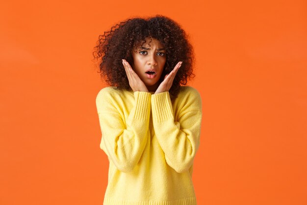 Chica encantadora tímida e insegura asustada con corte de pelo afro, suéter amarillo, jadeando sorprendida y preocupada, tocando y mirando con pánico, sintiendo miedo, de pie en la pared naranja.