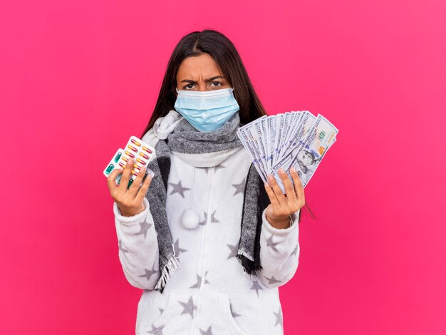 Chica enferma joven enojada con máscara médica con bufanda sosteniendo pastillas con efectivo aislado en rosa