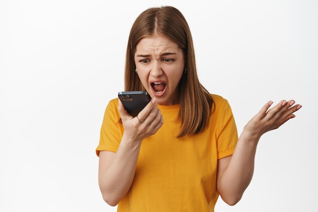 Chica enojada gritando en el mensaje de voz del teléfono inteligente, hablando en el altavoz y gritando en la dinámica del teléfono móvil, estrechando la mano frustrada, de pie contra el fondo blanco.