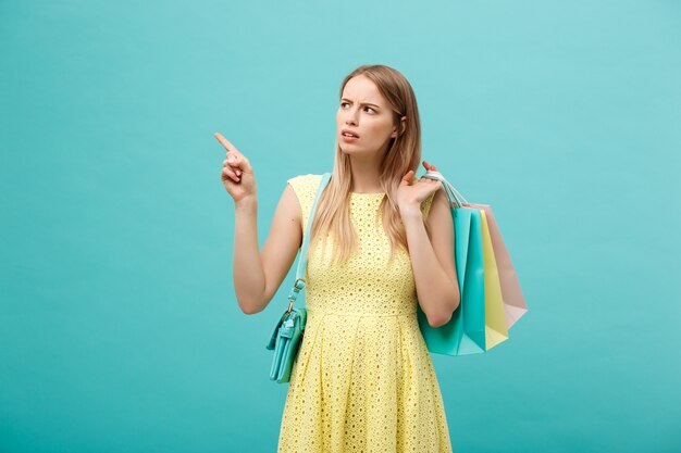 Chica enojada en vestido aislado sobre fondo azul. Sosteniendo la bolsa de papel de la compra para llevar y señalar con el dedo.