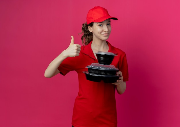 Chica de entrega bonita joven complacida con uniforme rojo y gorra sosteniendo contenedores de comida y mostrando el pulgar hacia arriba aislado sobre fondo carmesí con espacio de copia