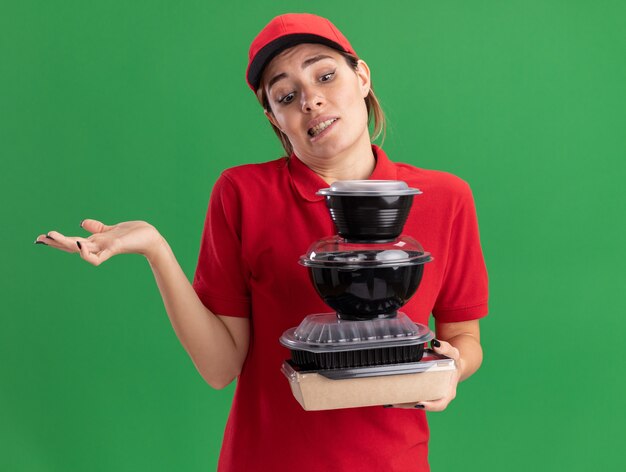 Chica de entrega bonita joven confundida en uniforme sostiene y mira los envases de comida en el paquete de comida en verde