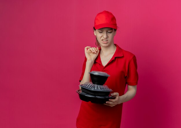 Chica de entrega bonita joven molesta con uniforme rojo y gorra sosteniendo y mirando contenedores de comida aislados sobre fondo carmesí con espacio de copia