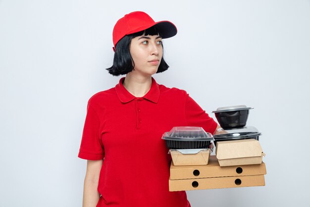 Chica de entrega caucásica joven complacida sosteniendo envases de alimentos y envases en cajas de pizza mirando al lado