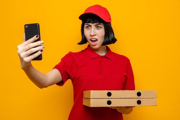 Chica de entrega caucásica joven molesta sosteniendo cajas de pizza y gritando a alguien mirando el teléfono