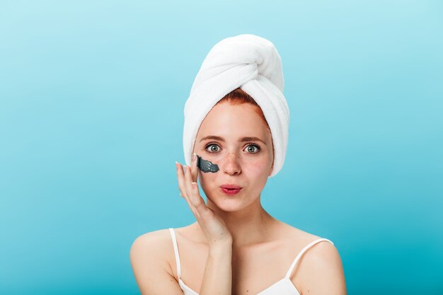 Chica entusiasta aplicando mascarilla. Disparo de estudio de mujer alegre haciendo tratamiento de spa y sonriendo sobre fondo azul.