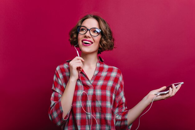 Chica entusiasta en auriculares posando con placer en la pared clarete. Foto interior de inspirada joven rizada lleva gafas y escucha música.