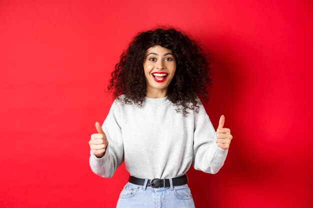 Chica entusiasta con cabello rizado y labios rojos, mostrando los pulgares hacia arriba y diciendo que sí, estoy de acuerdo contigo, felicita el buen trabajo, como algo genial, de pie en el fondo del estudio.