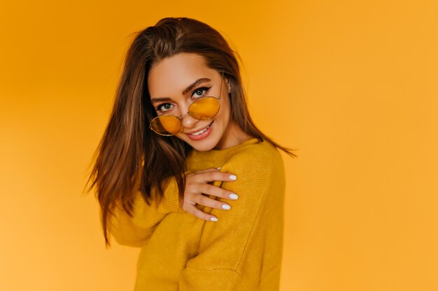 Chica entusiasta con manicura blanca posando con gafas de sol amarillas. Retrato de feliz dama europea con cabello castaño lacio.