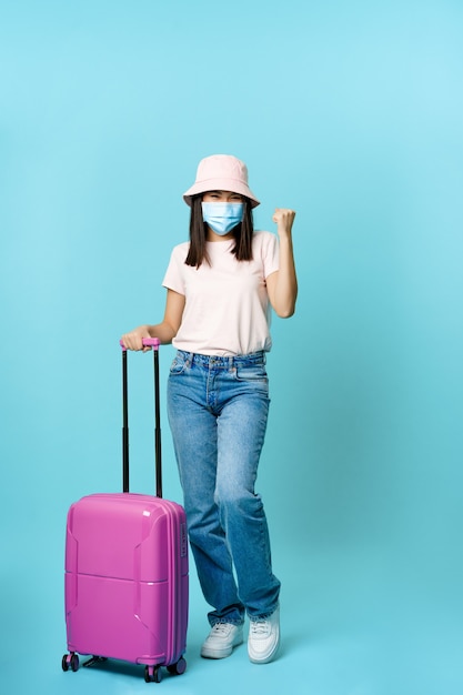 Chica entusiasta turista asiático en máscara médica sosteniendo la maleta tiro de cuerpo entero viajando al extranjero ...