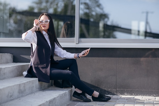 Chica en unas escaleras con un móvil en la mano y tocándose las gafas