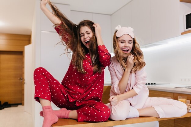 Chica escalofriante en calcetines rosas sentado en la mesa de madera. Foto interior de linda mujer joven en ropa de dormir rosa divirtiéndose en la cocina.