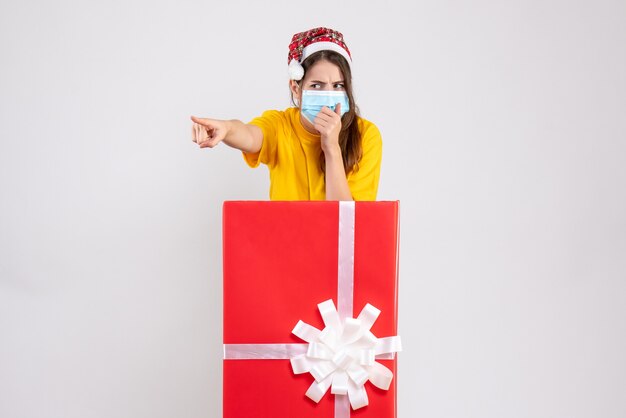 Chica escéptica con gorro de Papá Noel apuntando a algo detrás de un gran regalo de Navidad en blanco