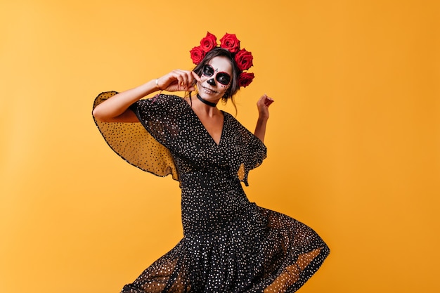 Chica española en vestido de gasa negro bailando danza folclórica y sonriendo. Foto de cuerpo entero de mujer con arte facial y rosas rojas en el pelo.