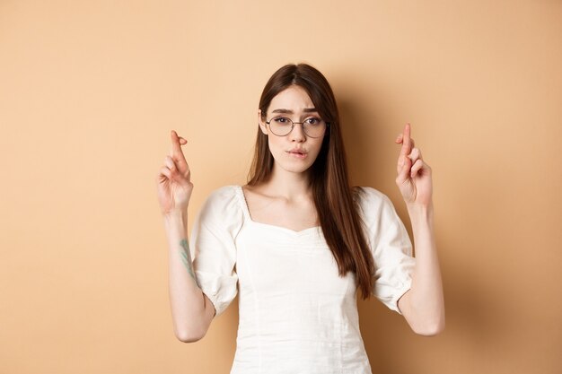 Chica esperanzada en gafas cruzar los dedos para la buena suerte mordiéndose el labio nervioso y mirando a la cámara esperando ...