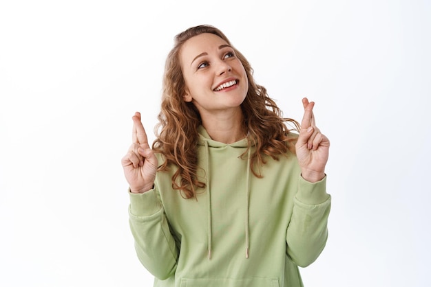 Una chica esperanzada con el pelo rubio y rizado cruza los dedos para tener buena suerte sonriendo y mirando el texto promocional rezando pidiendo un deseo de pie sobre un fondo blanco