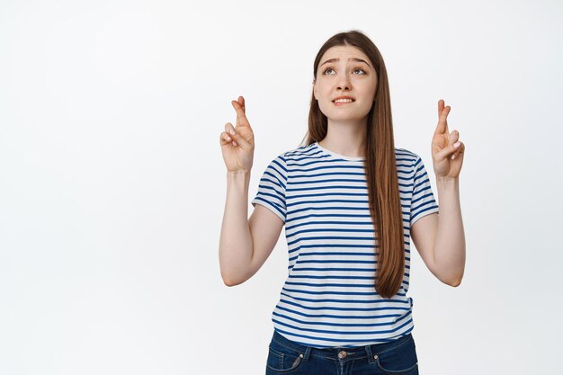 La chica esperanzada reza, cruza los dedos y mira hacia arriba nerviosamente, pidiendo un deseo contra el fondo blanco.