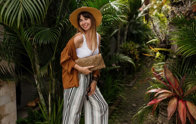 Chica con estilo elegante en top blanco y sombrero de paja posando en hojas de palma en Bali.
