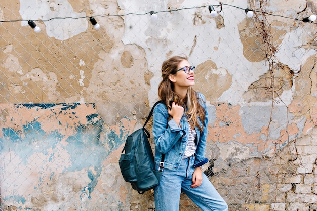 Chica con estilo hipster en el traje de jeans retro posando delante de la pared de ladrillo antiguo. Mujer joven de moda con pie de bolsa junto al edificio antiguo.