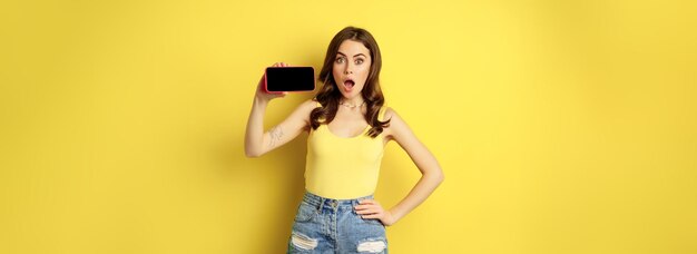 Chica con estilo sorprendida y sorprendida que muestra la aplicación de pantalla del teléfono inteligente en la pantalla horizontal del teléfono móvil