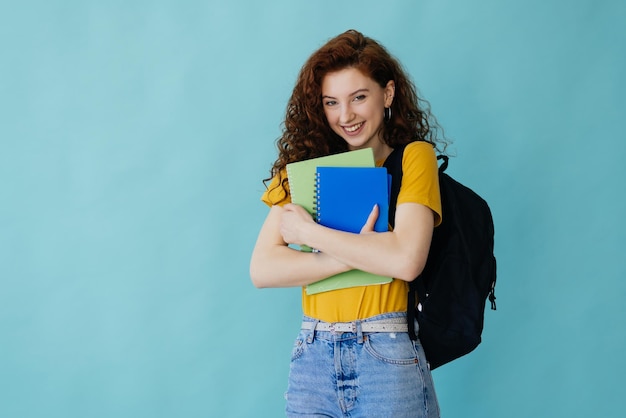 Chica estudiante adolescente aislada sobre fondo azul posando con los brazos en la cadera y sonriendo