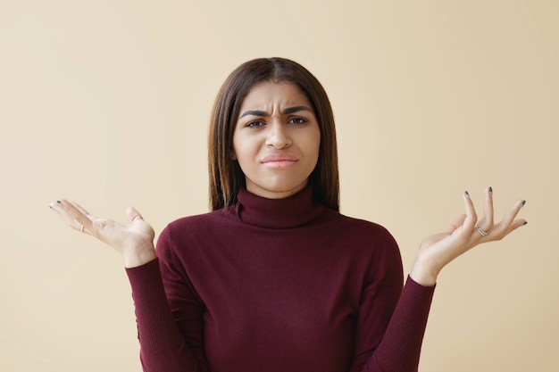 Chica estudiante africana disgustada expresando su indignación porque tiene que hacer muchos deberes. Fruncir el ceño joven mujer de raza mixta encogiéndose de hombros con las palmas abiertas, habiendo confundido mirada despistada