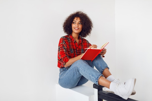 Chica estudiante africana pensativa descansando con el cuaderno. Con camisa roja a cuadros. Tejanos