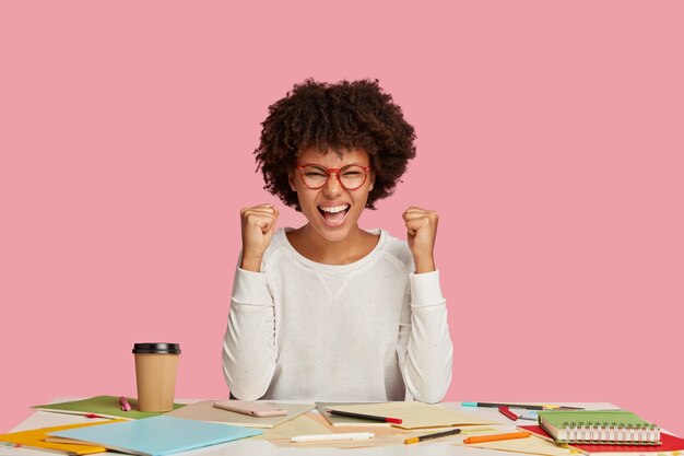 Chica estudiante alegre positiva posando en el escritorio contra la pared rosa