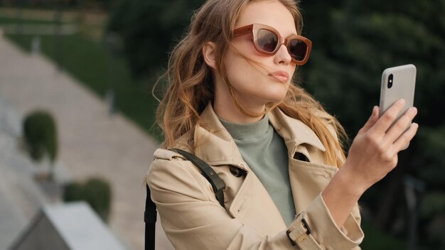 Chica estudiante atractiva con gafas de sol luciendo sensual tomando selfie para la red social en el teléfono inteligente en el parque de la ciudad