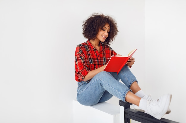 Chica estudiante bastante africana descansando con el cuaderno. Con camisa roja a cuadros. Tejanos