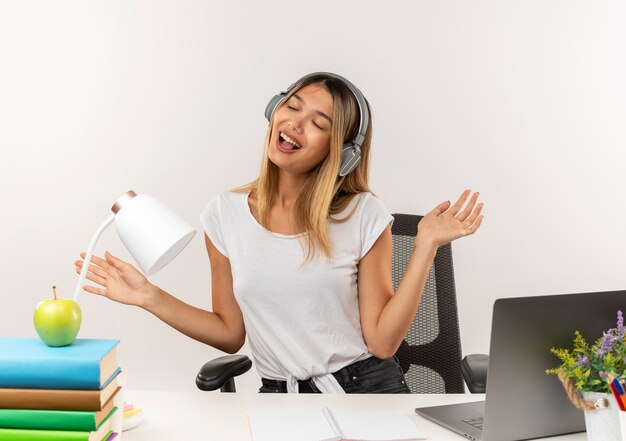 Chica estudiante bonita joven alegre con audífonos sentado en el escritorio con herramientas escolares escuchando música mostrando las manos vacías con los ojos cerrados aislados en blanco