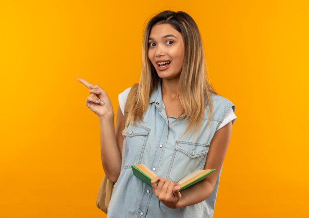 Chica estudiante bonita joven alegre con bolsa trasera sosteniendo libro abierto y apuntando al lado aislado en naranja