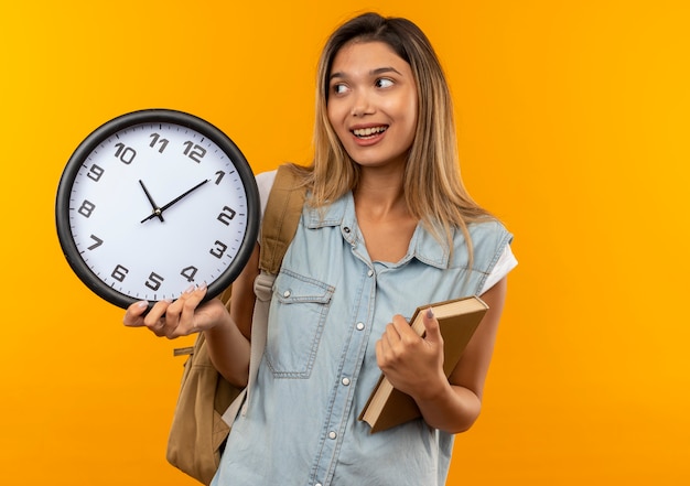 Chica estudiante bonita joven alegre con bolsa trasera sosteniendo libro y reloj y mirando al lado aislado en naranja