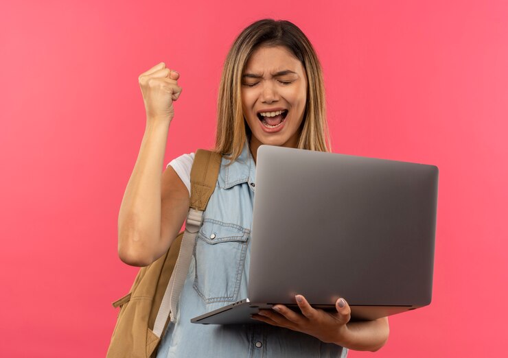 Foto gratuito chica estudiante bonita joven alegre con bolsa trasera sosteniendo portátil y levantando el puño con los ojos cerrados aislado en rosa