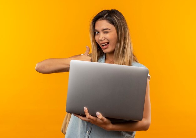 Chica estudiante bonita joven alegre con bolsa trasera sosteniendo portátil y mostrando el pulgar hacia arriba y guiñando un ojo aislado en naranja