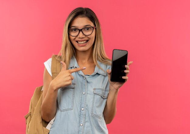 Chica estudiante bonita joven alegre con gafas y bolsa trasera sosteniendo y apuntando al teléfono móvil aislado en rosa