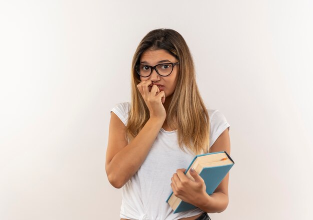 Chica estudiante bonita joven ansiosa con gafas y bolsa trasera sosteniendo el libro poniendo la mano en la barbilla aislada en blanco con espacio de copia