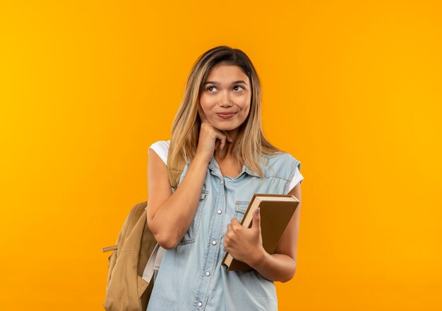 Chica estudiante bonita joven complacida con bolsa trasera sosteniendo el libro poniendo la mano debajo de la barbilla y mirando al lado aislado en la pared naranja