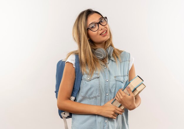 Chica estudiante bonita joven complacida con gafas y bolsa trasera con auriculares en el cuello sosteniendo libros aislados en la pared blanca