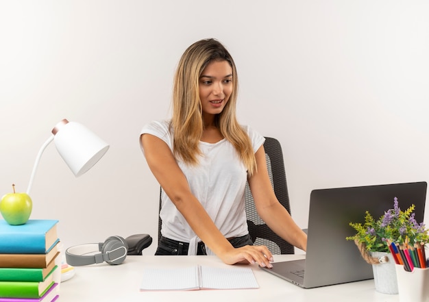 Chica estudiante bonita joven complacida de pie detrás del escritorio con herramientas escolares y usando laptop aislado en la pared blanca