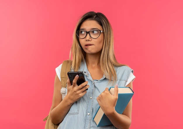 Chica estudiante bonita joven pensativa con gafas y bolso trasero sosteniendo teléfono móvil y libro mirando de lado y mordiendo el labio aislado en la pared rosa