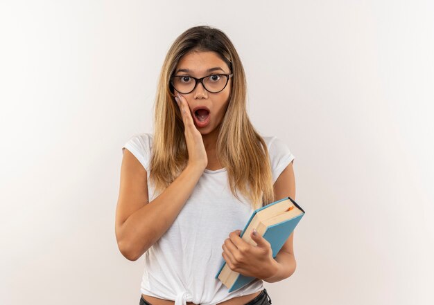 Chica estudiante bonita joven sorprendida con gafas y bolsa trasera sosteniendo el libro poniendo la mano en la mejilla aislada en la pared blanca